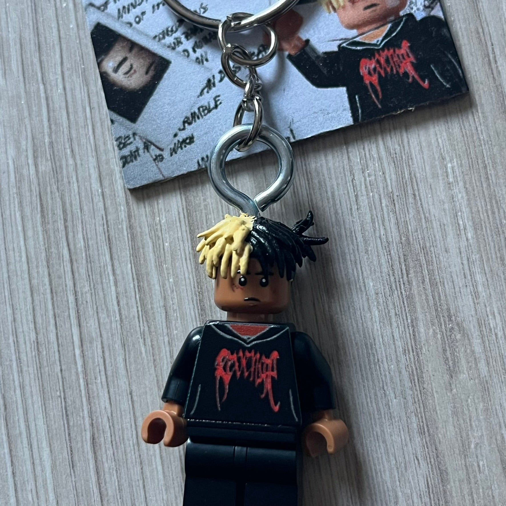 Lego XXXTentacion “XXXBlocktentacion” keychain featuring a custom Lego figure of XXXTentacion in a black/red revenge hoodie, with his classic split black and beige dreads in 3D. The figure is attached to a Lego-style recreation of the 2017 '17' album cover, reimagined in brick graphics. A great tribute and a creative collectible that merges music culture with fan-made Lego artistry, made by TheCanvasDon.