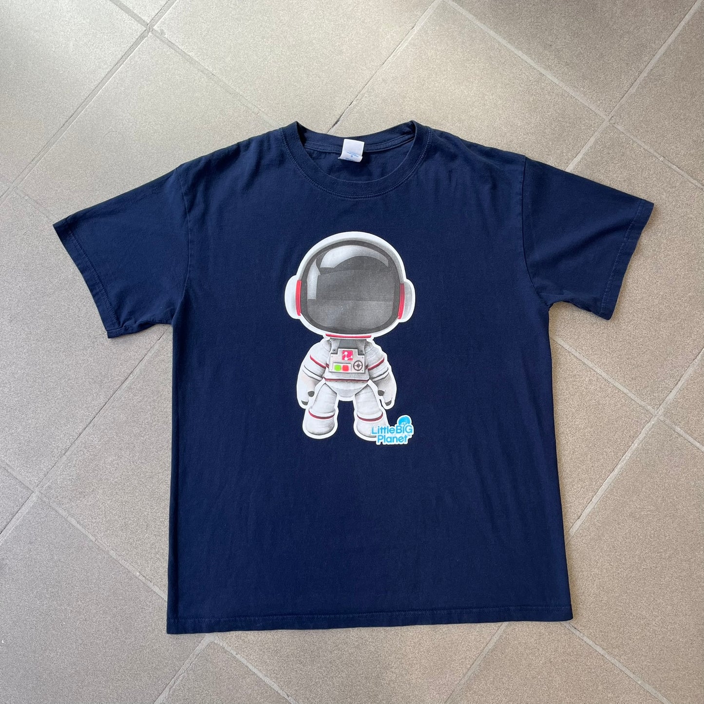 LittleBigPlanet 2011 Astronaut Navy Tee features a navy T-shirt made on a Gildan blank. The front has a white Sackboy graphic wearing an astronaut suit with a grey visor, accompanied by a blue LittleBigPlanet logo near the leg.
