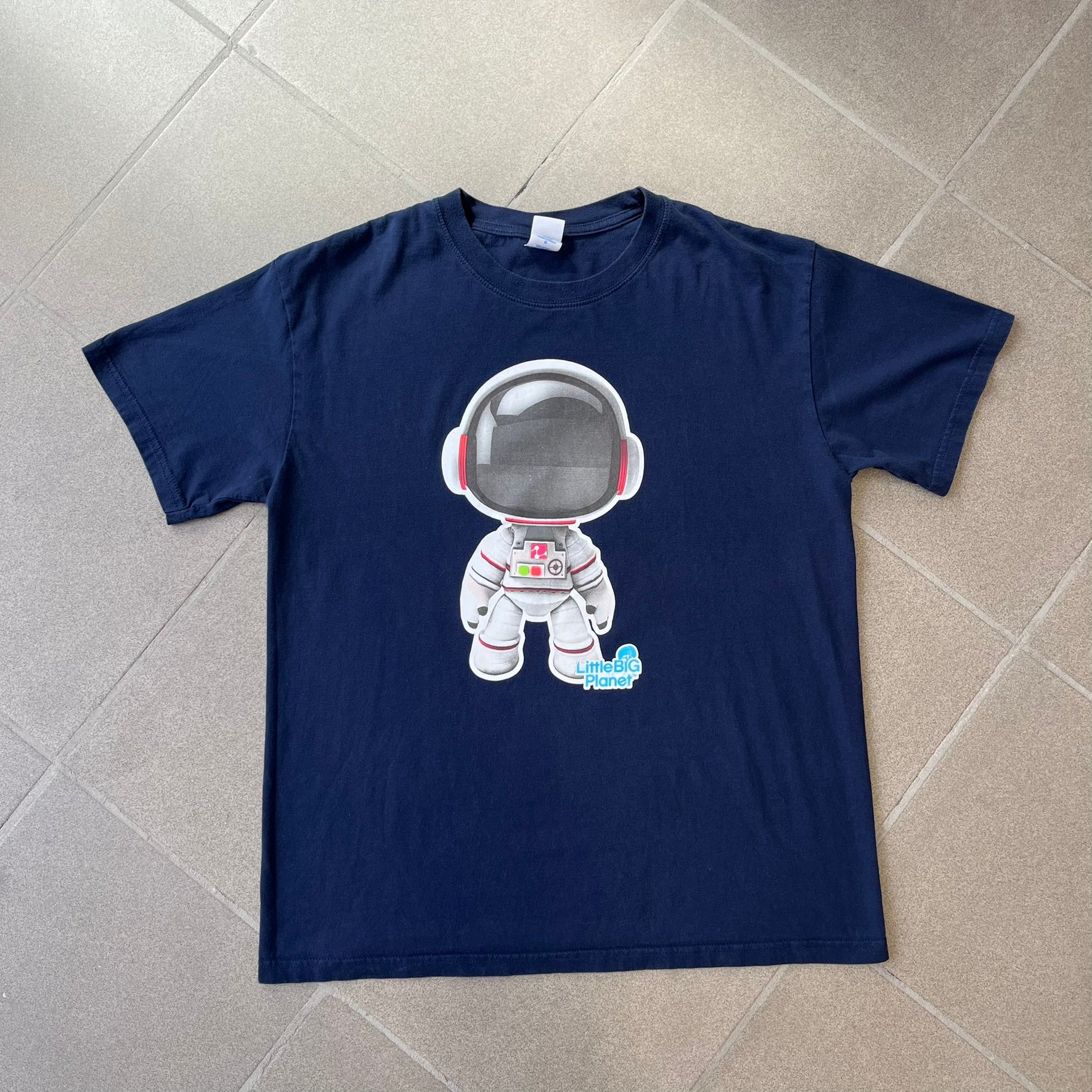 LittleBigPlanet 2011 Astronaut Navy Tee features a navy T-shirt made on a Gildan blank. The front has a white Sackboy graphic wearing an astronaut suit with a grey visor, accompanied by a blue LittleBigPlanet logo near the leg.
