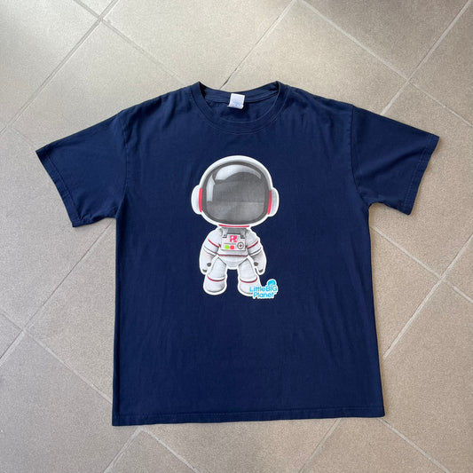 LittleBigPlanet 2011 Astronaut Navy Tee features a navy T-shirt made on a Gildan blank. The front has a white Sackboy graphic wearing an astronaut suit with a grey visor, accompanied by a blue LittleBigPlanet logo near the leg.
