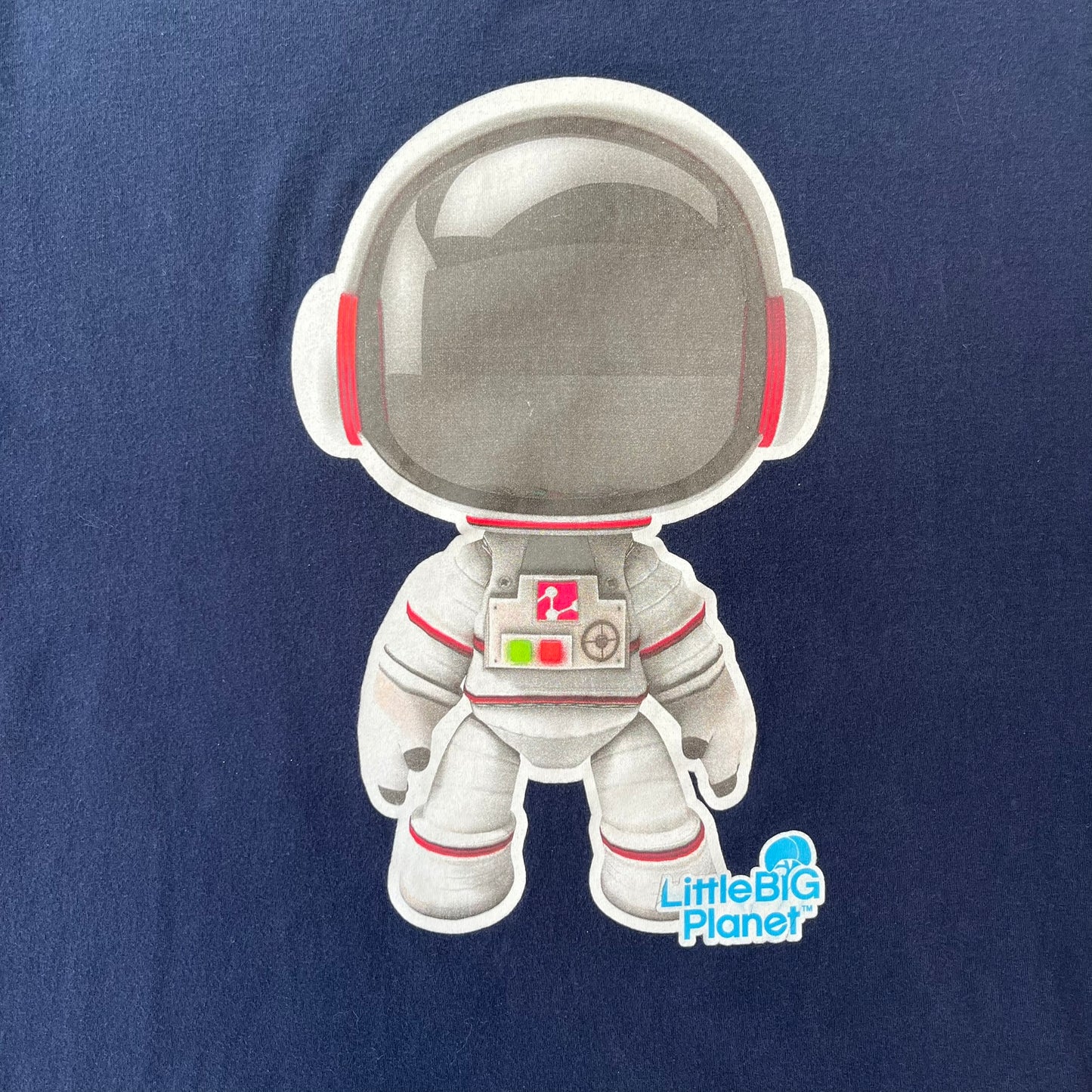LittleBigPlanet 2011 Astronaut Navy Tee features a navy T-shirt made on a Gildan blank. The front has a white Sackboy graphic wearing an astronaut suit with a grey visor, accompanied by a blue LittleBigPlanet logo near the leg.
