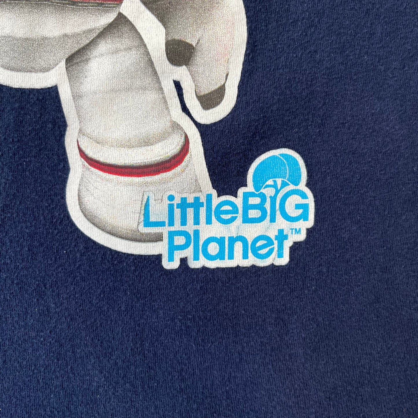 LittleBigPlanet 2011 Astronaut Navy Tee features a navy T-shirt made on a Gildan blank. The front has a white Sackboy graphic wearing an astronaut suit with a grey visor, accompanied by a blue LittleBigPlanet logo near the leg.
