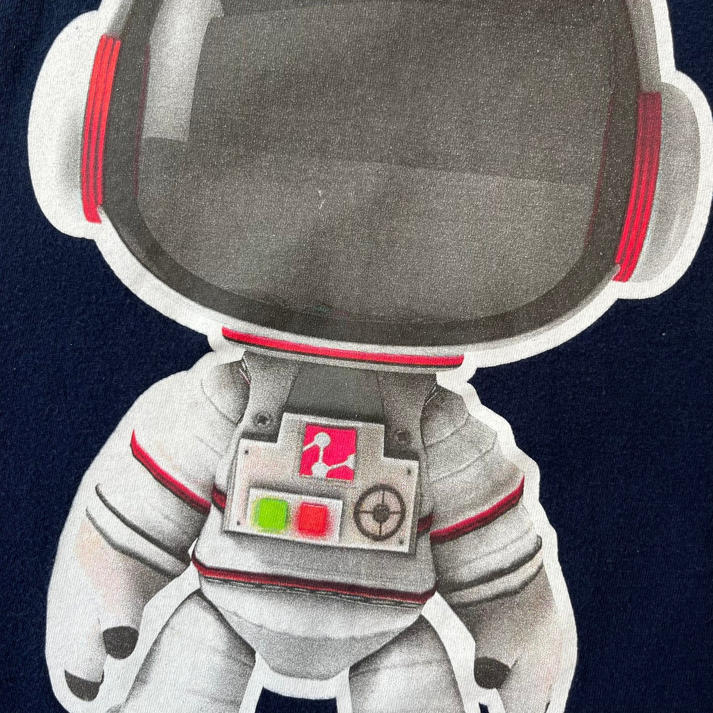LittleBigPlanet 2011 Astronaut Navy Tee features a navy T-shirt made on a Gildan blank. The front has a white Sackboy graphic wearing an astronaut suit with a grey visor, accompanied by a blue LittleBigPlanet logo near the leg.
