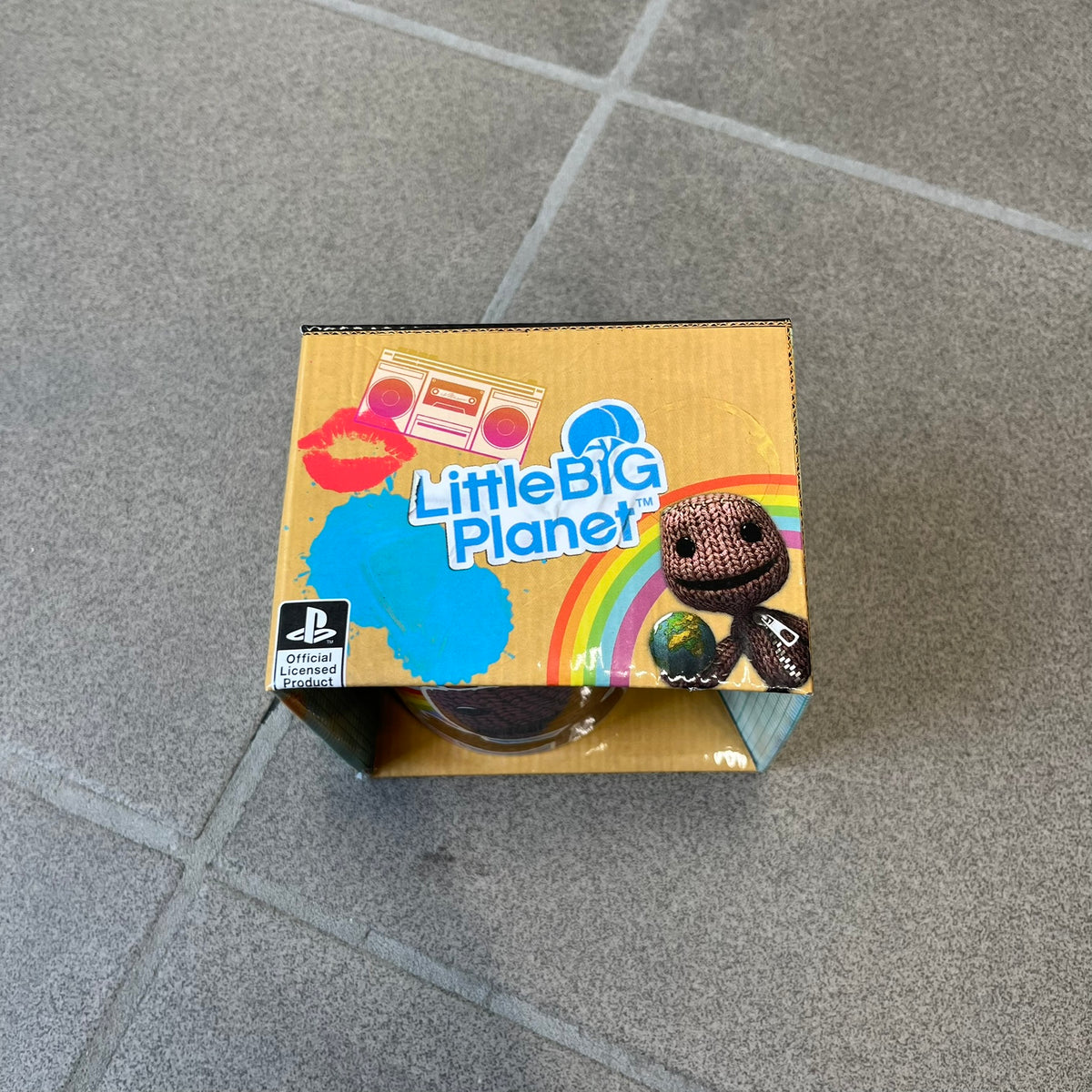 Little Big Planet 2010 Collectable Mug is a white mug with a blue wraparound graphic of a happy Sackboy, the LittleBigPlanet logo with “Play, Create, Share” text, and other LBP graphics set against a paint splatter and sticker-style background. It is officially licensed by Sony Computer Entertainment, with SCEE 2010 licensing on the bottom. It comes in its original box, which has multiple graphics and licensing.&nbsp;