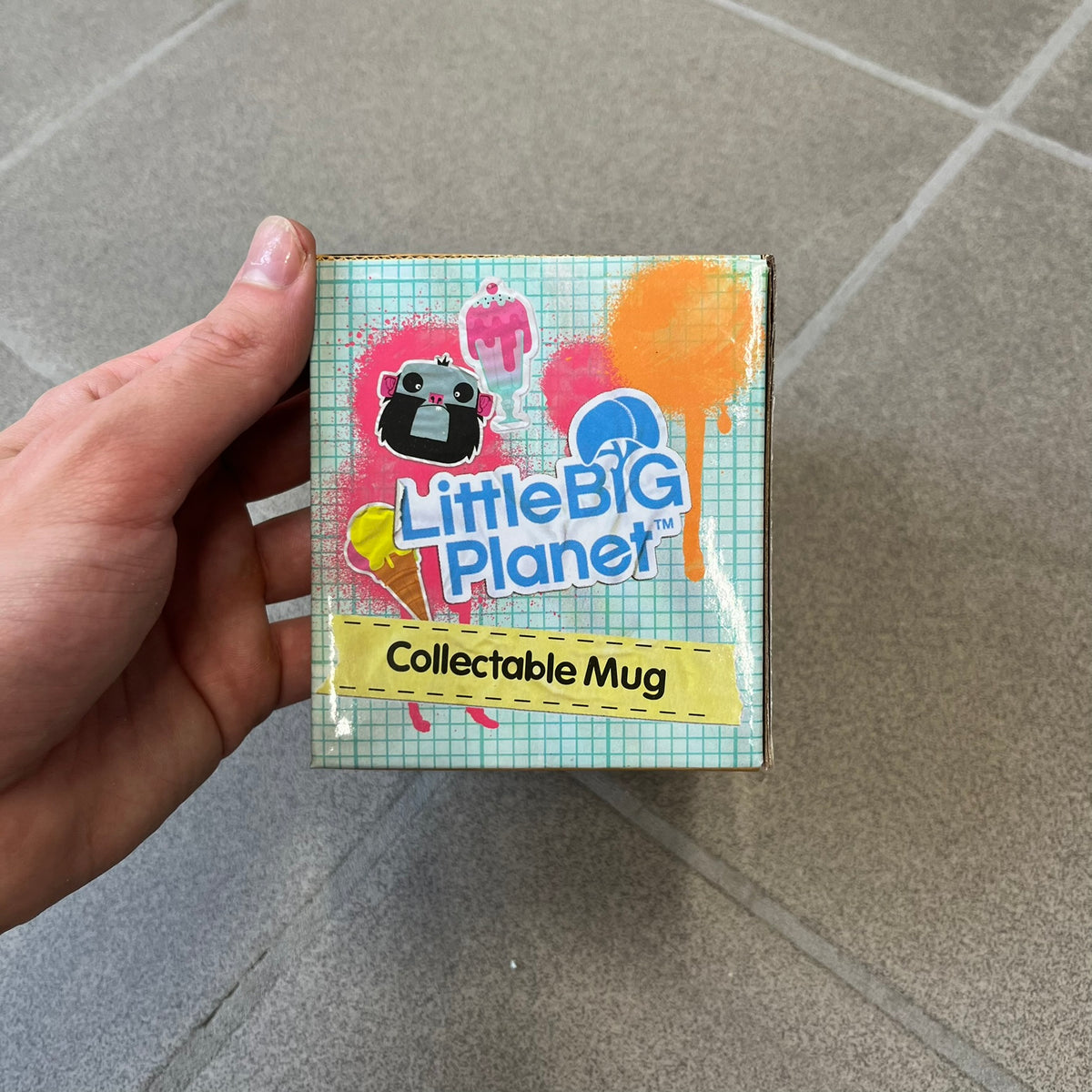 Little Big Planet 2010 Collectable Mug is a white mug with a blue wraparound graphic of a happy Sackboy, the LittleBigPlanet logo with “Play, Create, Share” text, and other LBP graphics set against a paint splatter and sticker-style background. It is officially licensed by Sony Computer Entertainment, with SCEE 2010 licensing on the bottom. It comes in its original box, which has multiple graphics and licensing.&nbsp;