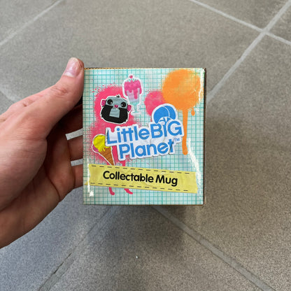 Little Big Planet 2010 Collectable Mug is a white mug with a blue wraparound graphic of a happy Sackboy, the LittleBigPlanet logo with “Play, Create, Share” text, and other LBP graphics set against a paint splatter and sticker-style background. It is officially licensed by Sony Computer Entertainment, with SCEE 2010 licensing on the bottom. It comes in its original box, which has multiple graphics and licensing.&nbsp;