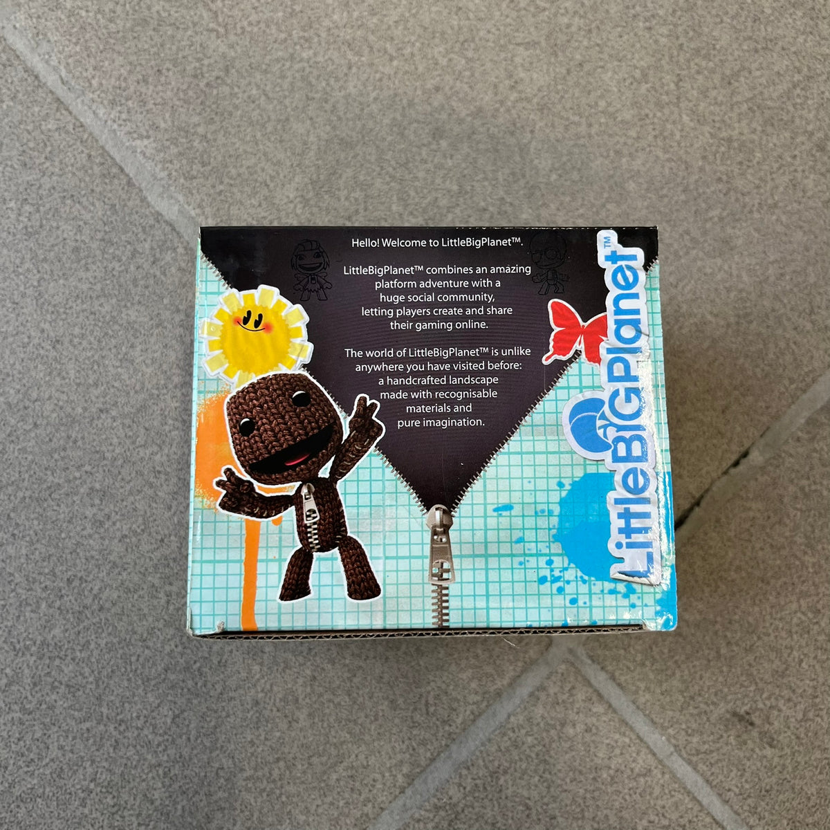 Little Big Planet 2010 Collectable Mug is a white mug with a blue wraparound graphic of a happy Sackboy, the LittleBigPlanet logo with “Play, Create, Share” text, and other LBP graphics set against a paint splatter and sticker-style background. It is officially licensed by Sony Computer Entertainment, with SCEE 2010 licensing on the bottom. It comes in its original box, which has multiple graphics and licensing.&nbsp;