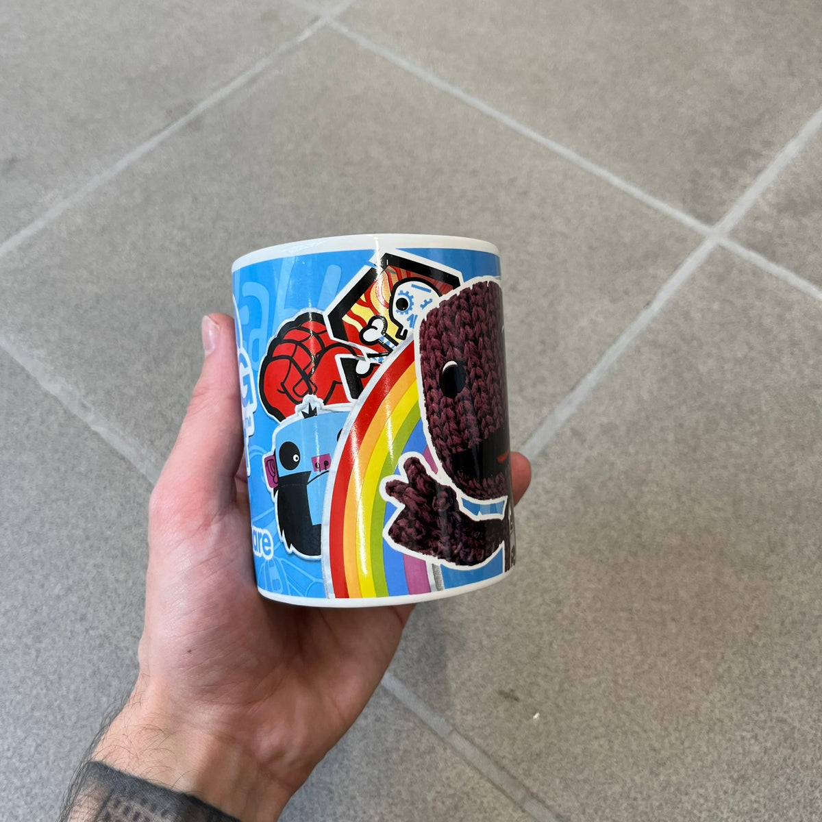 Little Big Planet 2010 Collectable Mug is a white mug with a blue wraparound graphic of a happy Sackboy, the LittleBigPlanet logo with “Play, Create, Share” text, and other LBP graphics set against a paint splatter and sticker-style background. It is officially licensed by Sony Computer Entertainment, with SCEE 2010 licensing on the bottom. It comes in its original box, which has multiple graphics and licensing.&nbsp;