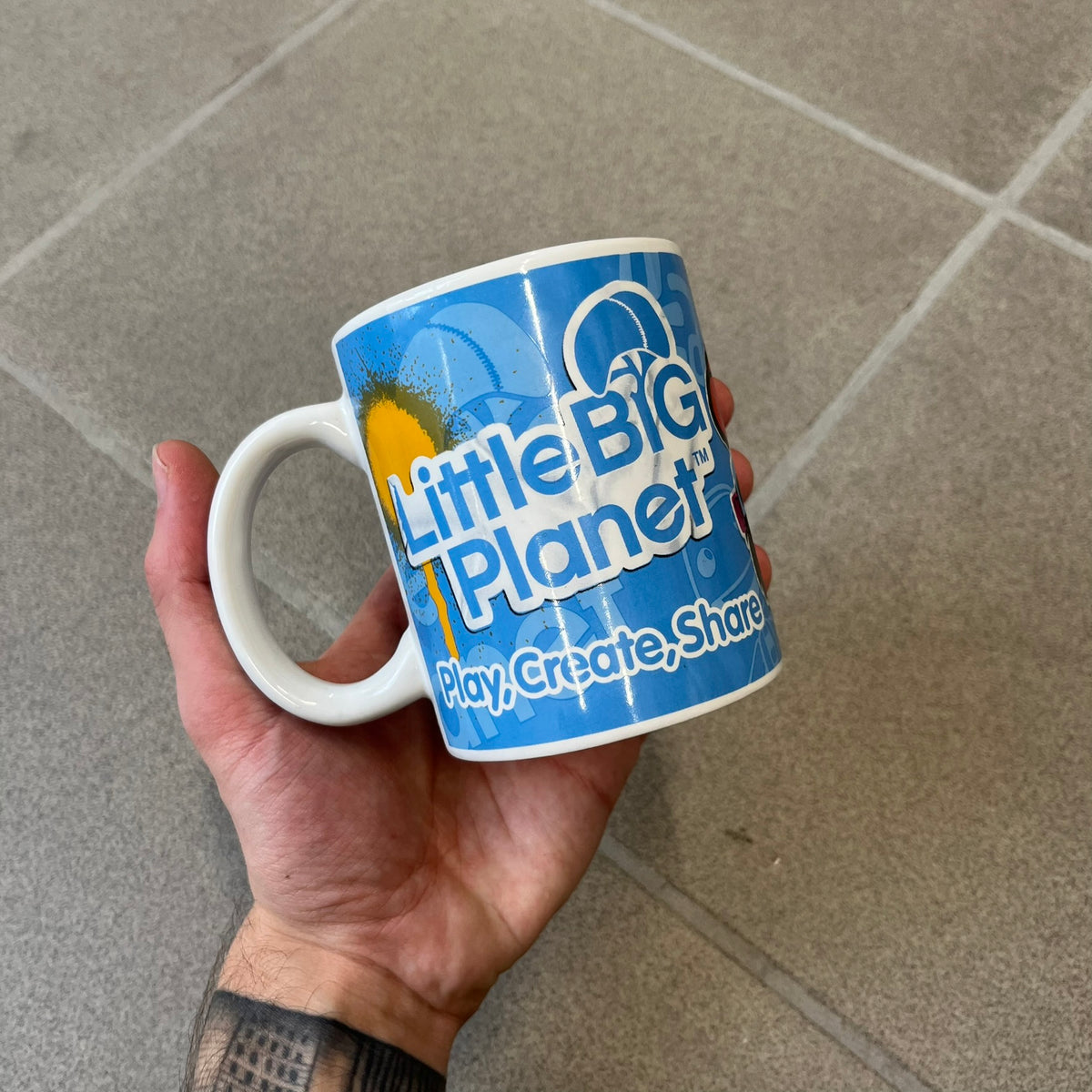 Little Big Planet 2010 Collectable Mug is a white mug with a blue wraparound graphic of a happy Sackboy, the LittleBigPlanet logo with “Play, Create, Share” text, and other LBP graphics set against a paint splatter and sticker-style background. It is officially licensed by Sony Computer Entertainment, with SCEE 2010 licensing on the bottom. It comes in its original box, which has multiple graphics and licensing.&nbsp;
