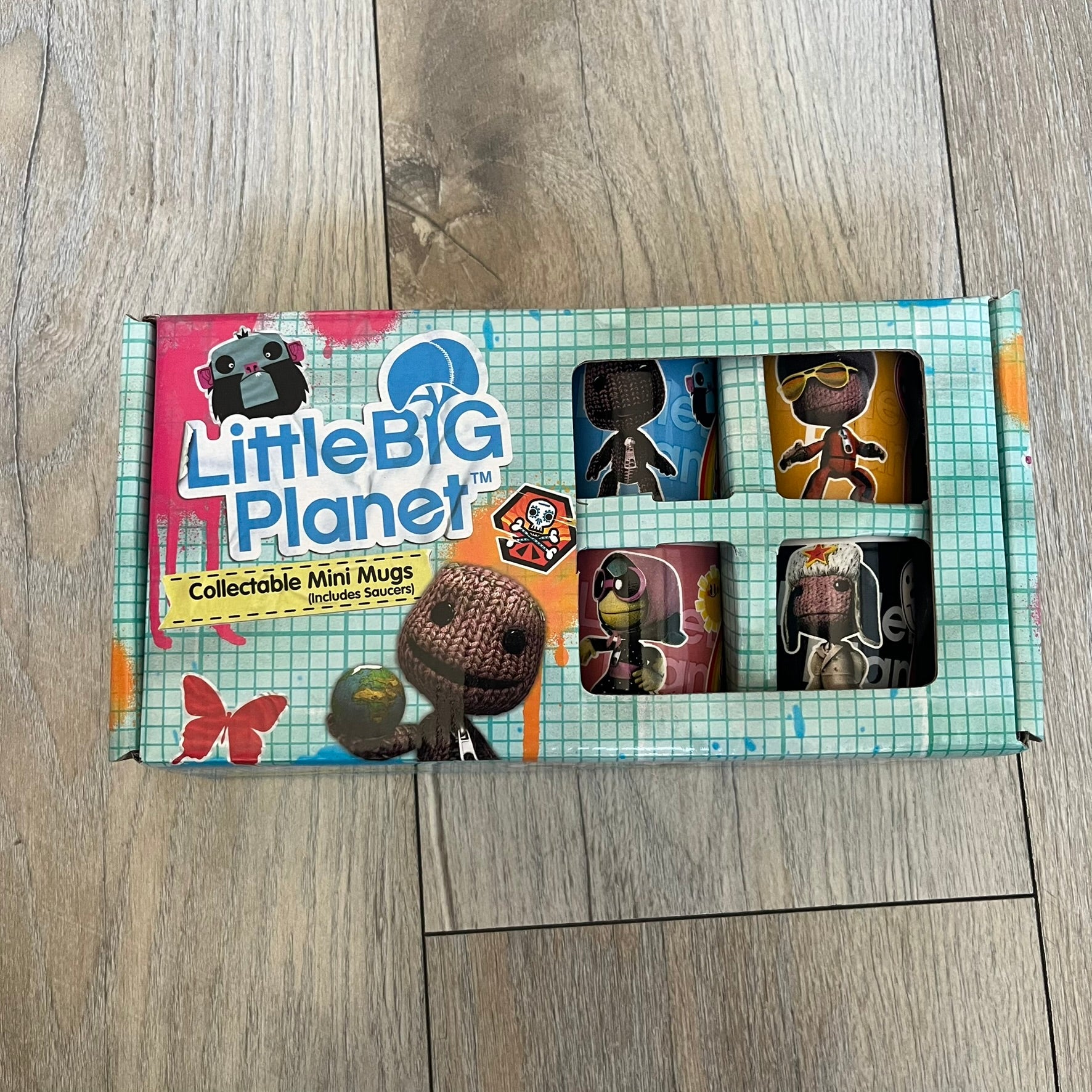 LittleBigPlanet 2011 Collectable Mini Mugs + Saucers Set is a 4-piece espresso set featuring characters from the LittleBigPlanet series, including Sackboy, Marvin the Disco Dancer, Gloria, and Yuri Army Sackboy. Each mini mug includes a wraparound character graphic with the LittleBigPlanet logo and comes with a matching saucer printed on the top. The set is officially licensed by Sony Computer Entertainment, includes a display box, and carries SCEE 2011 licensing on the base of the mugs and saucers.