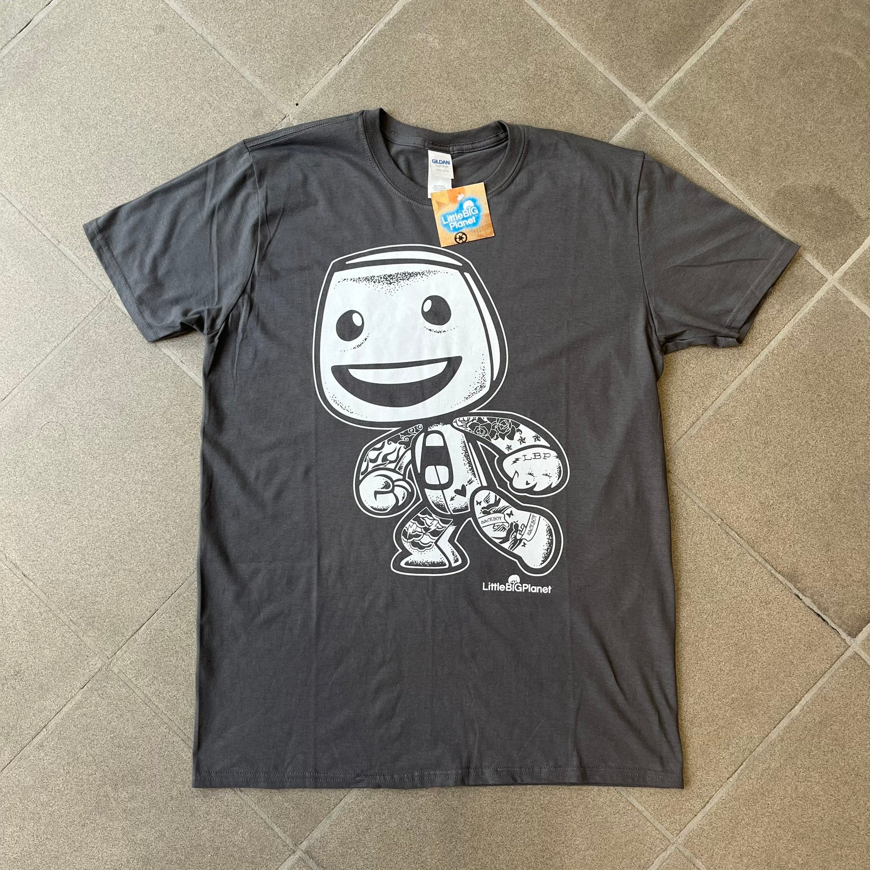 LittleBigPlanet 2016 Tattoo Sackboy Tee features a grey T-shirt made on a Gildan blank. The front has a large white graphic of a running Sackboy covered in tattoo designs, with a LittleBigPlanet logo underneath. Includes the original paper LittleBigPlanet tag.

