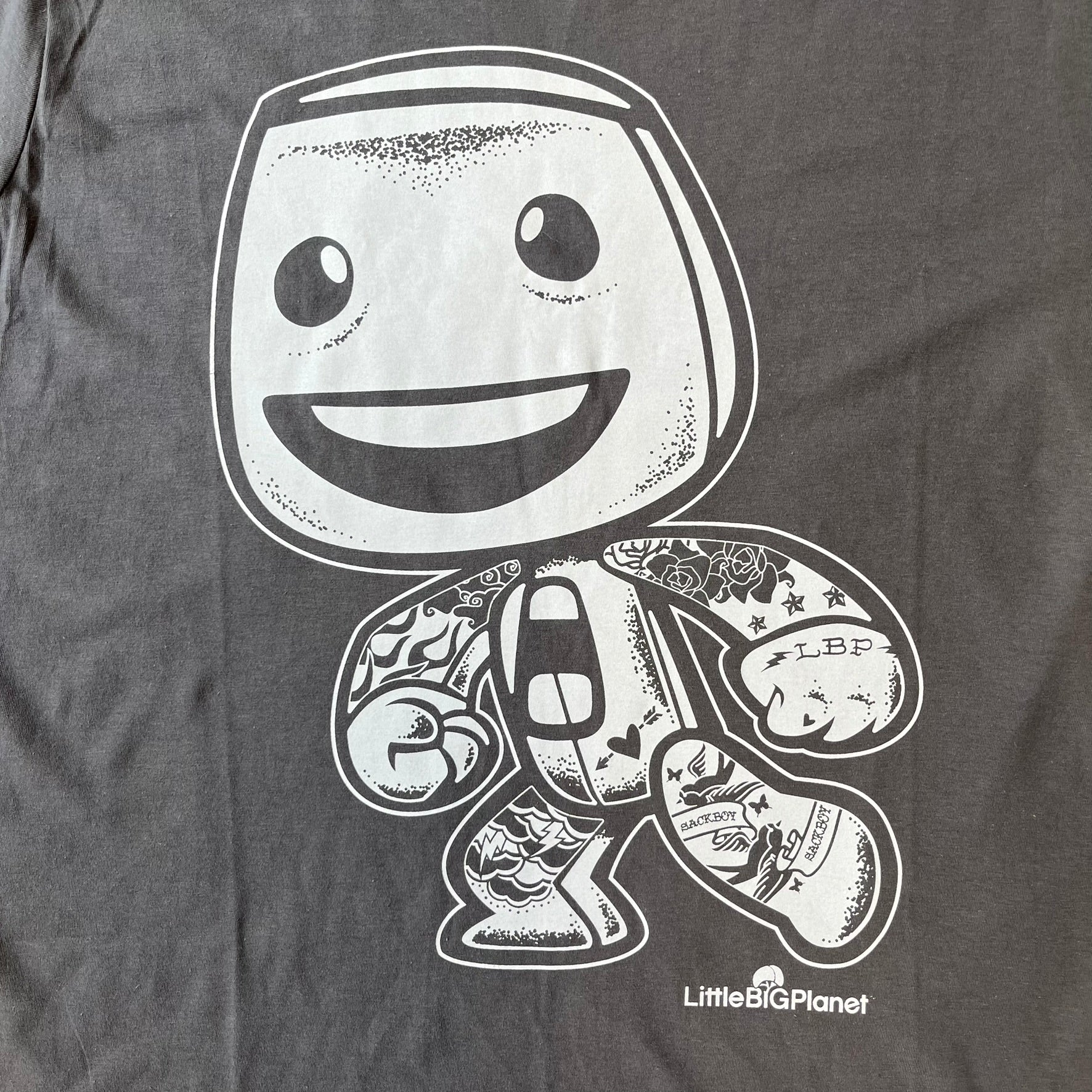 LittleBigPlanet 2016 Tattoo Sackboy Tee features a grey T-shirt made on a Gildan blank. The front has a large white graphic of a running Sackboy covered in tattoo designs, with a LittleBigPlanet logo underneath. Includes the original paper LittleBigPlanet tag.
