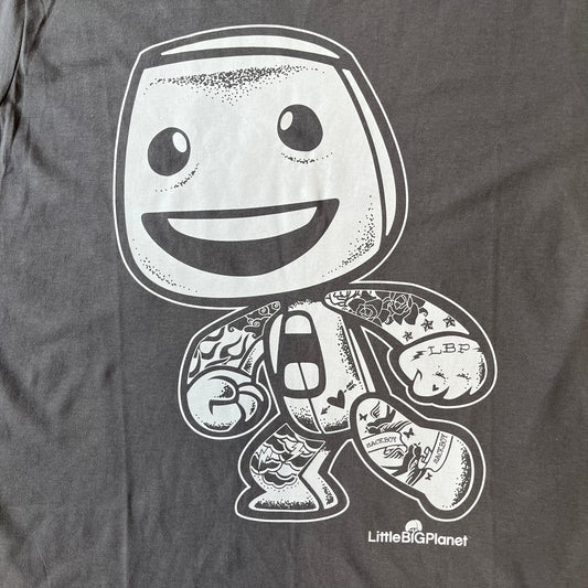 LittleBigPlanet 2016 Tattoo Sackboy Tee features a grey T-shirt made on a Gildan blank. The front has a large white graphic of a running Sackboy covered in tattoo designs, with a LittleBigPlanet logo underneath. Includes the original paper LittleBigPlanet tag.
