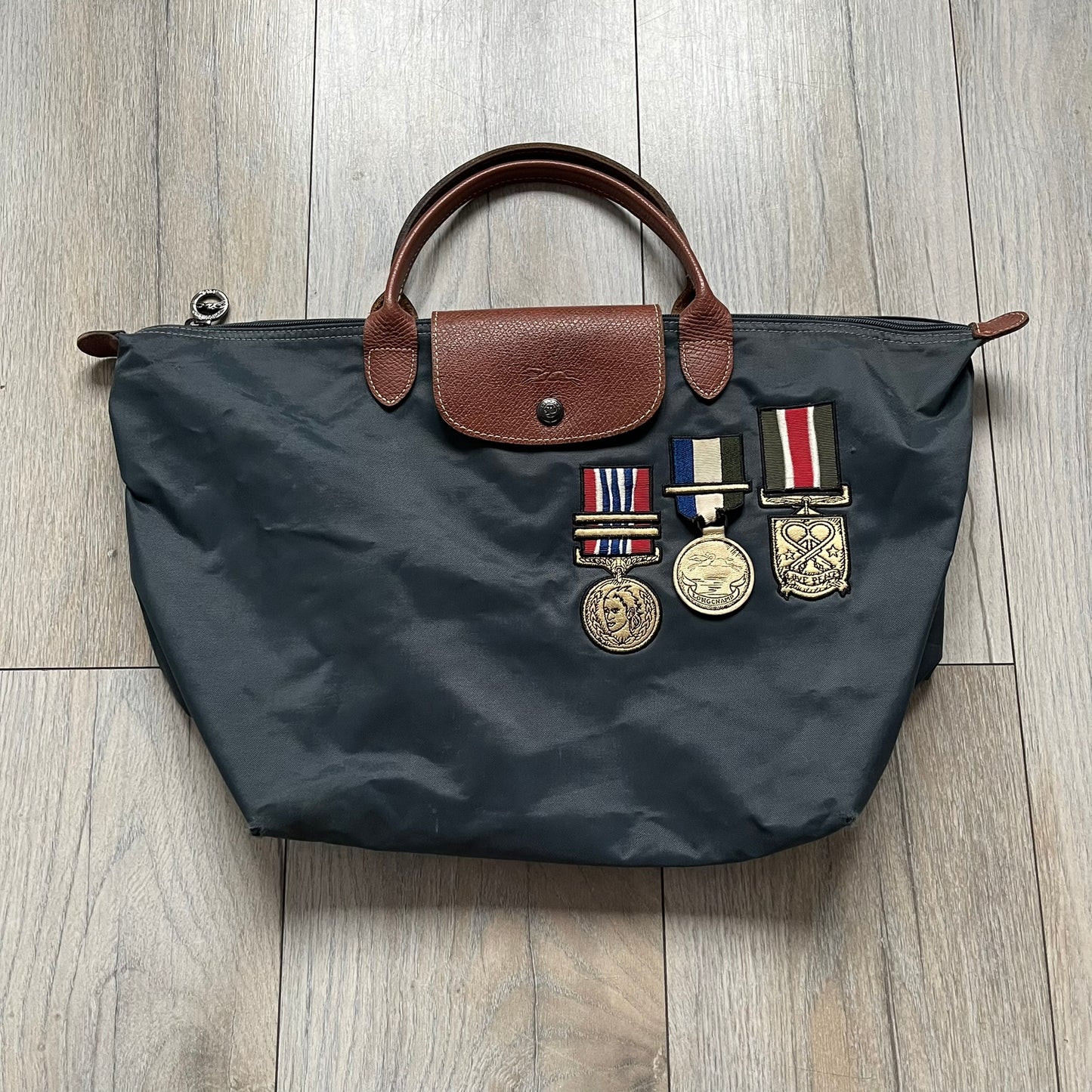 Longchamp x Jeremy Scott 2009 “Medals” Le Pliage Large Handbag is a grey nylon foldable tote with brown leather handles and flap, featuring a branded metal zipper and an interior Jeremy Scott x Longchamp leather tag, made in France. The front showcases embroidered medal motifs, including a Jeremy Scott portrait, Longchamp logo, and key emblem, with one medal designed as a movable attachment. This design is part of the A/W 2009 Military-inspired Colonel line collaboration.