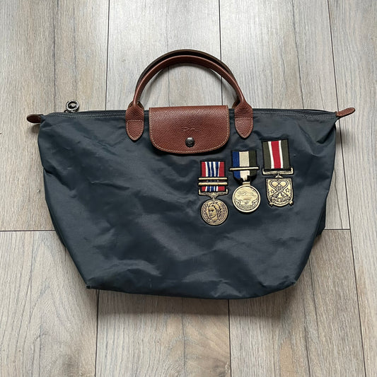 Longchamp x Jeremy Scott 2009 “Medals” Le Pliage Large Handbag is a grey nylon foldable tote with brown leather handles and flap, featuring a branded metal zipper and an interior Jeremy Scott x Longchamp leather tag, made in France. The front showcases embroidered medal motifs, including a Jeremy Scott portrait, Longchamp logo, and key emblem, with one medal designed as a movable attachment. This design is part of the A/W 2009 Military-inspired Colonel line collaboration.