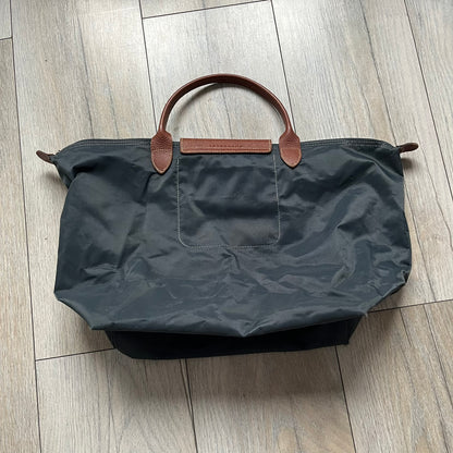 Longchamp x Jeremy Scott 2009 “Medals” Le Pliage Large Handbag is a grey nylon foldable tote with brown leather handles and flap, featuring a branded metal zipper and an interior Jeremy Scott x Longchamp leather tag, made in France. The front showcases embroidered medal motifs, including a Jeremy Scott portrait, Longchamp logo, and key emblem, with one medal designed as a movable attachment. This design is part of the A/W 2009 Military-inspired Colonel line collaboration.