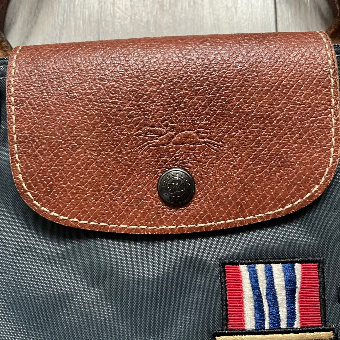 Longchamp x Jeremy Scott 2009 “Medals” Le Pliage Large Handbag is a grey nylon foldable tote with brown leather handles and flap, featuring a branded metal zipper and an interior Jeremy Scott x Longchamp leather tag, made in France. The front showcases embroidered medal motifs, including a Jeremy Scott portrait, Longchamp logo, and key emblem, with one medal designed as a movable attachment. This design is part of the A/W 2009 Military-inspired Colonel line collaboration.