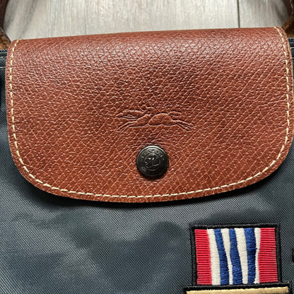 Longchamp x Jeremy Scott 2009 “Medals” Le Pliage Large Handbag is a grey nylon foldable tote with brown leather handles and flap, featuring a branded metal zipper and an interior Jeremy Scott x Longchamp leather tag, made in France. The front showcases embroidered medal motifs, including a Jeremy Scott portrait, Longchamp logo, and key emblem, with one medal designed as a movable attachment. This design is part of the A/W 2009 Military-inspired Colonel line collaboration.