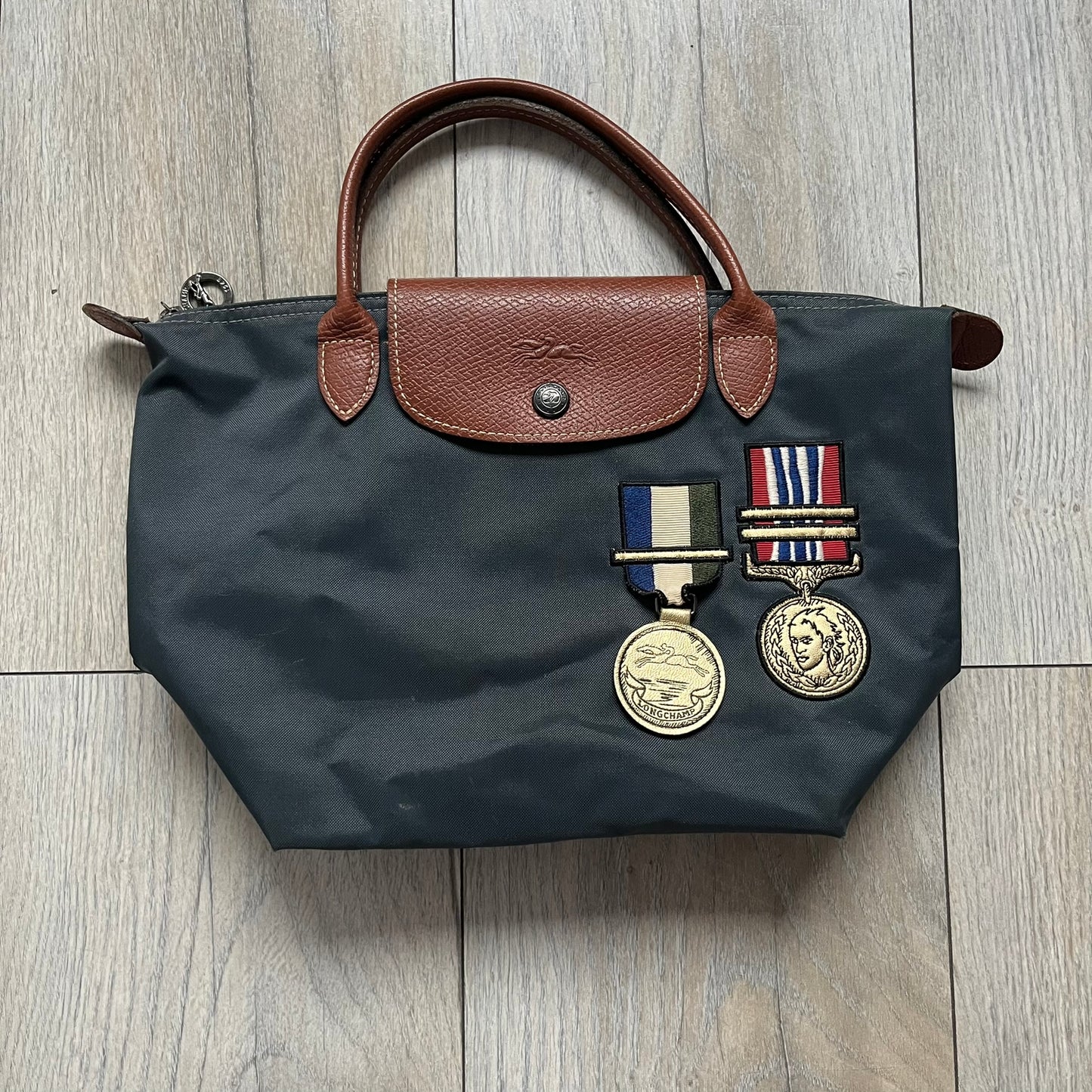 Longchamp x Jeremy Scott 2009 “Medals” Le Pliage Small Handbag is a grey nylon foldable tote with brown leather handles and flap, featuring a branded metal zipper and an interior Jeremy Scott x Longchamp leather tag, made in France. The front displays embroidered medal motifs, including a Jeremy Scott portrait and Longchamp logo, with one medal designed as a movable attachment. This piece is part of the A/W 2009 Military-inspired Colonel line collaboration.