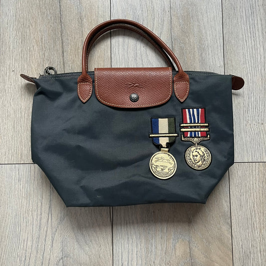 Longchamp x Jeremy Scott 2009 “Medals” Le Pliage Small Handbag is a grey nylon foldable tote with brown leather handles and flap, featuring a branded metal zipper and an interior Jeremy Scott x Longchamp leather tag, made in France. The front displays embroidered medal motifs, including a Jeremy Scott portrait and Longchamp logo, with one medal designed as a movable attachment. This piece is part of the A/W 2009 Military-inspired Colonel line collaboration.