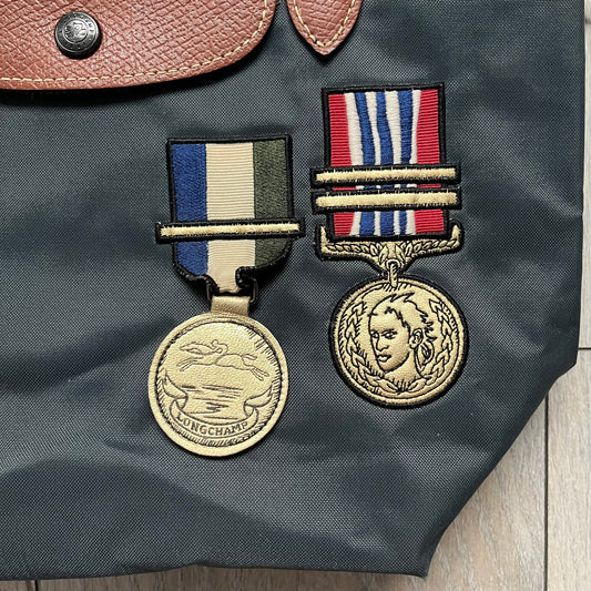 Longchamp x Jeremy Scott 2009 “Medals” Le Pliage Small Handbag is a grey nylon foldable tote with brown leather handles and flap, featuring a branded metal zipper and an interior Jeremy Scott x Longchamp leather tag, made in France. The front displays embroidered medal motifs, including a Jeremy Scott portrait and Longchamp logo, with one medal designed as a movable attachment. This piece is part of the A/W 2009 Military-inspired Colonel line collaboration.