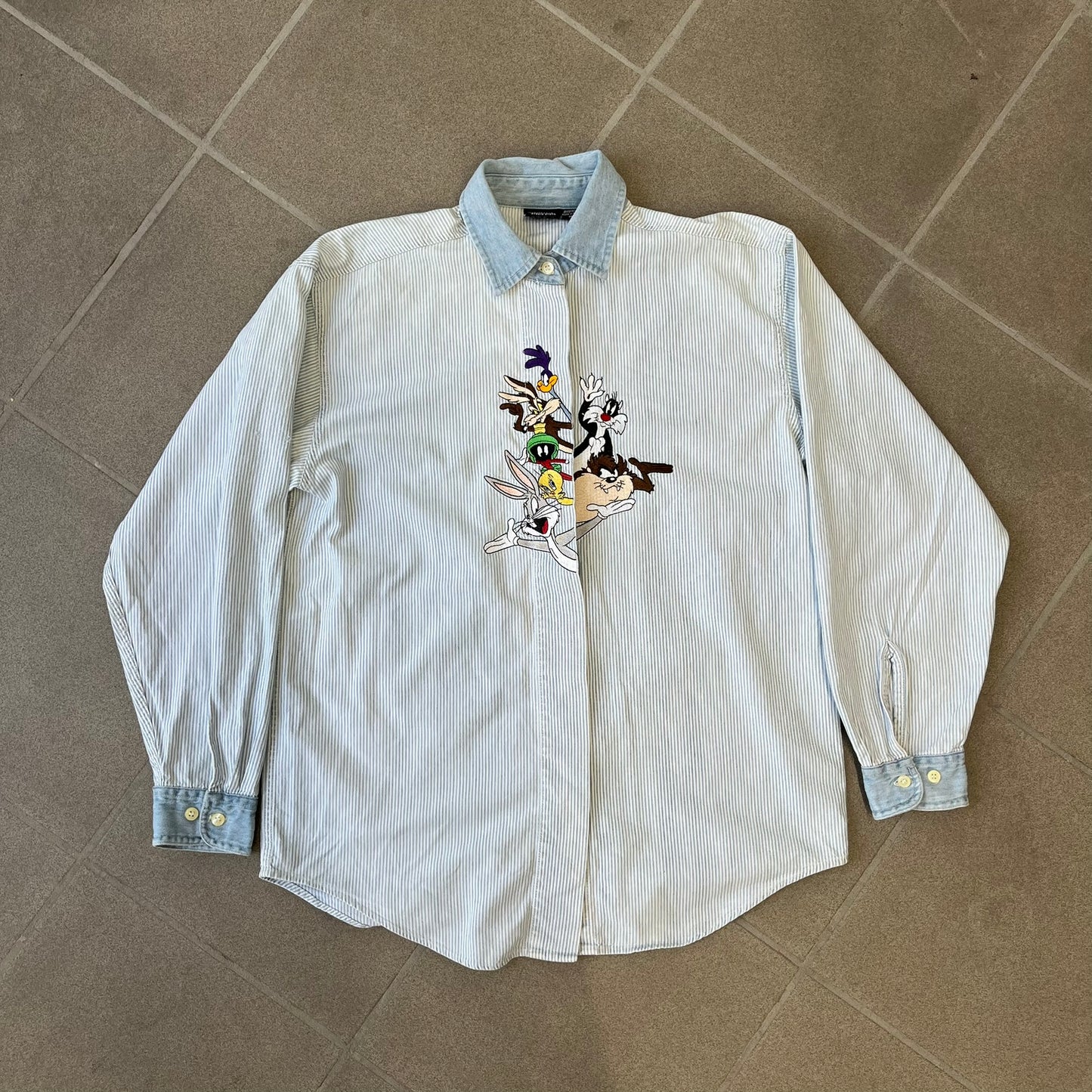 Looney Tunes 1996 Embroidered Striped Shirt features a light blue striped 100% cotton construction with a denim collar and matching denim hems. The center has a colorful embroidered design featuring the Looney Tunes characters Marsian, Bugs Bunny, Taz, Tweety, Sylvester, Road Runner, and Wile E. Coyote appearing from the middle. Made in Hong Kong and finished with a 1996 black licensed neck tag.