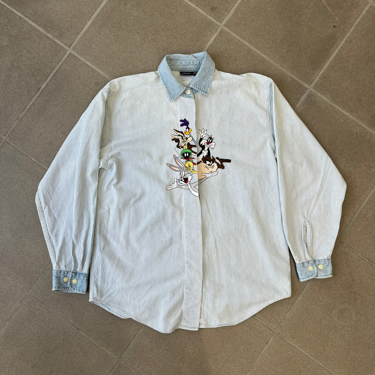 Looney Tunes 1996 Embroidered Striped Shirt features a light blue striped 100% cotton construction with a denim collar and matching denim hems. The center has a colorful embroidered design featuring the Looney Tunes characters Marsian, Bugs Bunny, Taz, Tweety, Sylvester, Road Runner, and Wile E. Coyote appearing from the middle. Made in Hong Kong and finished with a 1996 black licensed neck tag.