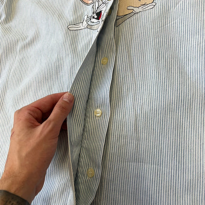 Looney Tunes 1996 Embroidered Striped Shirt features a light blue striped 100% cotton construction with a denim collar and matching denim hems. The center has a colorful embroidered design featuring the Looney Tunes characters Marsian, Bugs Bunny, Taz, Tweety, Sylvester, Road Runner, and Wile E. Coyote appearing from the middle. Made in Hong Kong and finished with a 1996 black licensed neck tag.