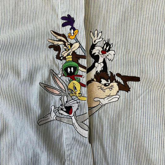 Looney Tunes 1996 Embroidered Striped Shirt features a light blue striped 100% cotton construction with a denim collar and matching denim hems. The center has a colorful embroidered design featuring the Looney Tunes characters Marsian, Bugs Bunny, Taz, Tweety, Sylvester, Road Runner, and Wile E. Coyote appearing from the middle. Made in Hong Kong and finished with a 1996 black licensed neck tag.