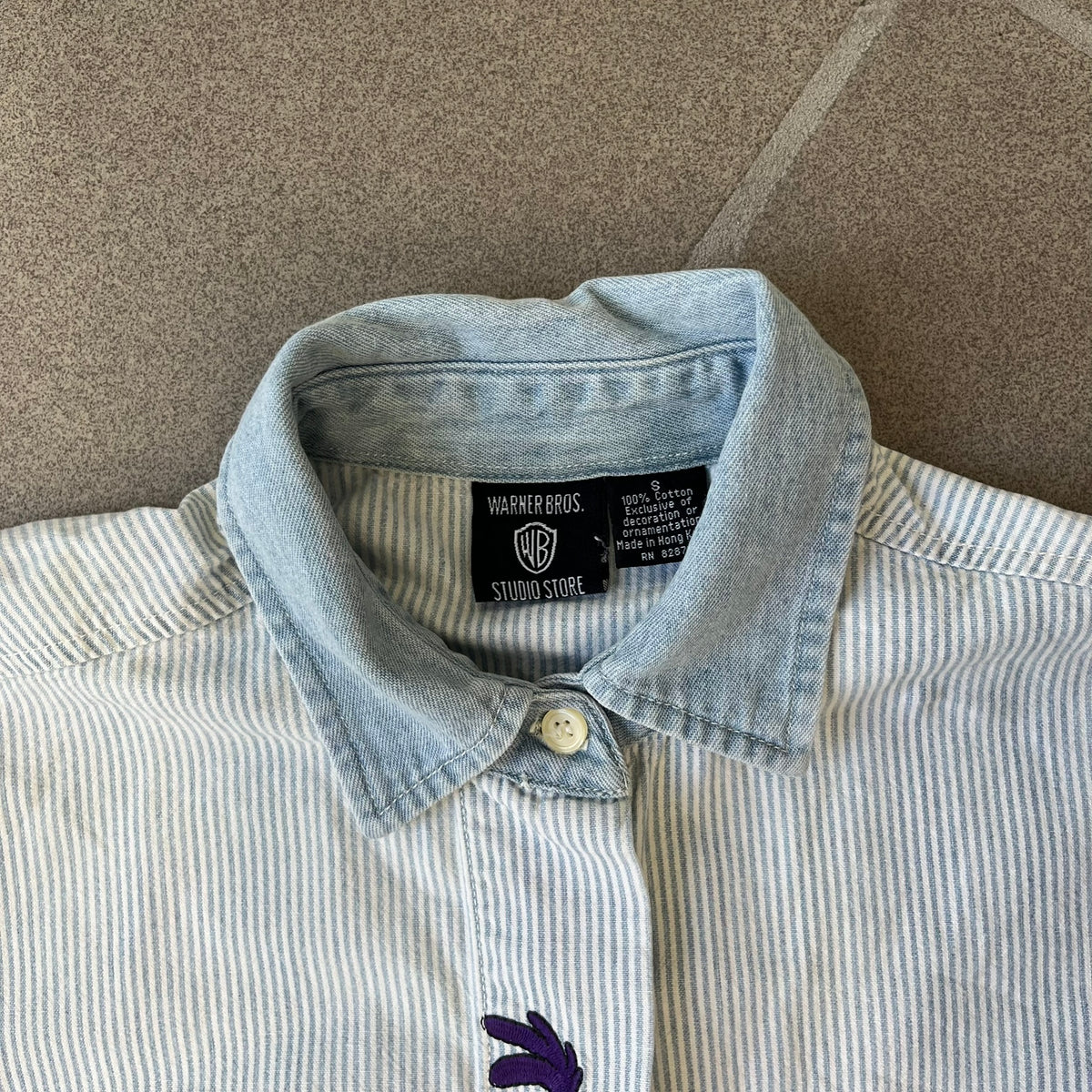 Looney Tunes 1996 Embroidered Striped Shirt features a light blue striped 100% cotton construction with a denim collar and matching denim hems. The center has a colorful embroidered design featuring the Looney Tunes characters Marsian, Bugs Bunny, Taz, Tweety, Sylvester, Road Runner, and Wile E. Coyote appearing from the middle. Made in Hong Kong and finished with a 1996 black licensed neck tag.