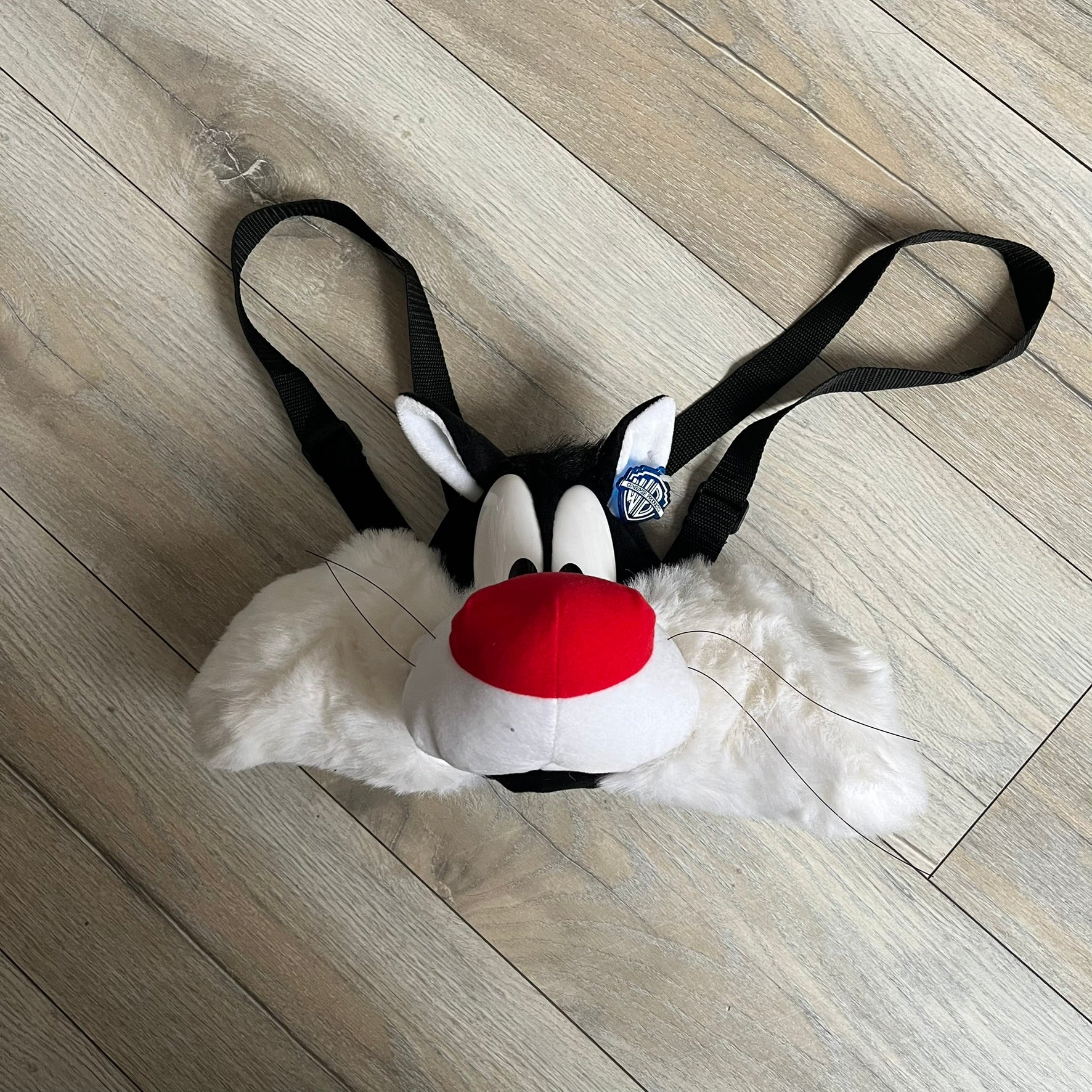 Looney Tunes 1996 Sylvester Head Backpack is a backpack shaped like Sylvester’s head. It features a furry exterior, white plastic eyes, a red nose, and detailed character features throughout. The backpack includes black shoulder straps, a blue plastic Warner Bros. tag, and a 1996 licensing tag.
