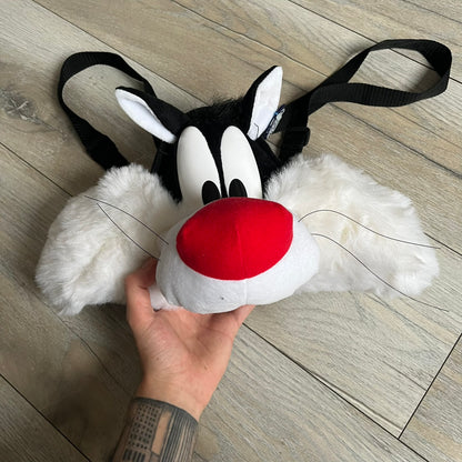 Looney Tunes 1996 Sylvester Head Backpack is a backpack shaped like Sylvester’s head. It features a furry exterior, white plastic eyes, a red nose, and detailed character features throughout. The backpack includes black shoulder straps, a blue plastic Warner Bros. tag, and a 1996 licensing tag.