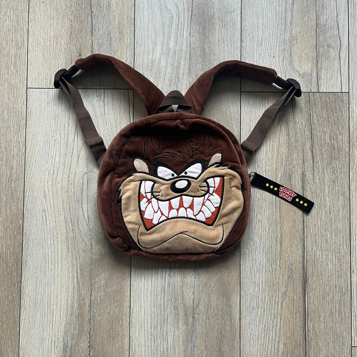 Small plush backpack made from soft brown material, featuring a large 3D Tasmanian Devil face on the front panel. Equipped with a zip closure detailed with a black Looney Tunes branded zipper pull, similar to a keychain tag. Finished with original Looney Tunes tags and compact backpack proportions suitable for light carry.