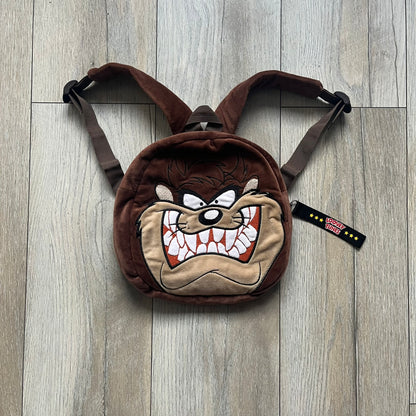 Small plush backpack made from soft brown material, featuring a large 3D Tasmanian Devil face on the front panel. Equipped with a zip closure detailed with a black Looney Tunes branded zipper pull, similar to a keychain tag. Finished with original Looney Tunes tags and compact backpack proportions suitable for light carry.