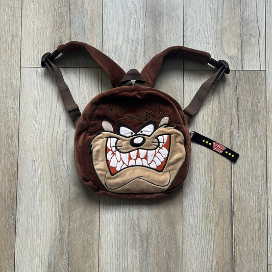 Small plush backpack made from soft brown material, featuring a large 3D Tasmanian Devil face on the front panel. Equipped with a zip closure detailed with a black Looney Tunes branded zipper pull, similar to a keychain tag. Finished with original Looney Tunes tags and compact backpack proportions suitable for light carry.