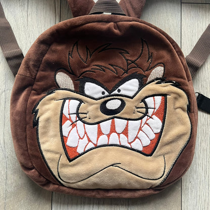 Small plush backpack made from soft brown material, featuring a large 3D Tasmanian Devil face on the front panel. Equipped with a zip closure detailed with a black Looney Tunes branded zipper pull, similar to a keychain tag. Finished with original Looney Tunes tags and compact backpack proportions suitable for light carry.