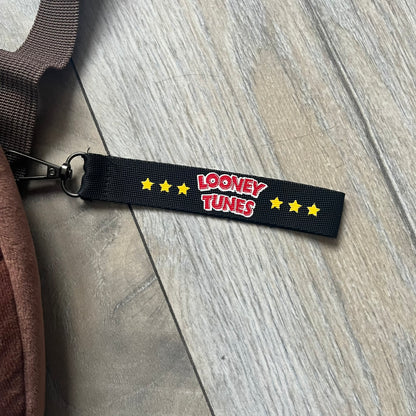 Small plush backpack made from soft brown material, featuring a large 3D Tasmanian Devil face on the front panel. Equipped with a zip closure detailed with a black Looney Tunes branded zipper pull, similar to a keychain tag. Finished with original Looney Tunes tags and compact backpack proportions suitable for light carry.
