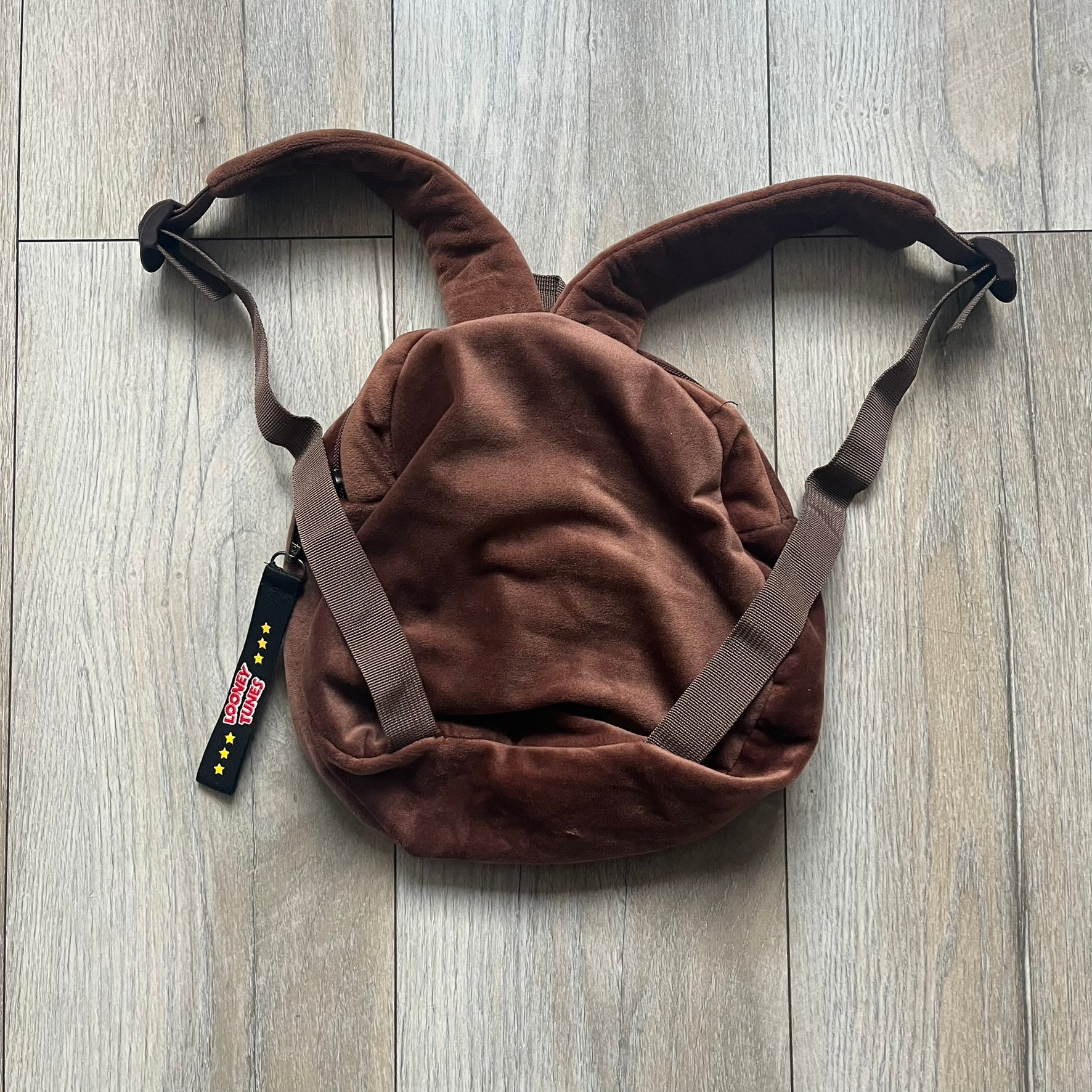 Small plush backpack made from soft brown material, featuring a large 3D Tasmanian Devil face on the front panel. Equipped with a zip closure detailed with a black Looney Tunes branded zipper pull, similar to a keychain tag. Finished with original Looney Tunes tags and compact backpack proportions suitable for light carry.
