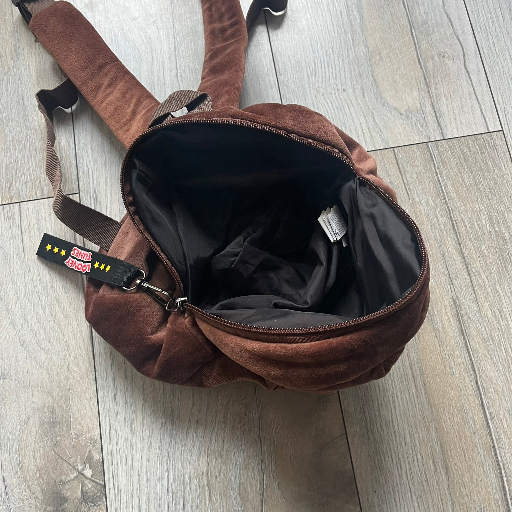 Small plush backpack made from soft brown material, featuring a large 3D Tasmanian Devil face on the front panel. Equipped with a zip closure detailed with a black Looney Tunes branded zipper pull, similar to a keychain tag. Finished with original Looney Tunes tags and compact backpack proportions suitable for light carry.