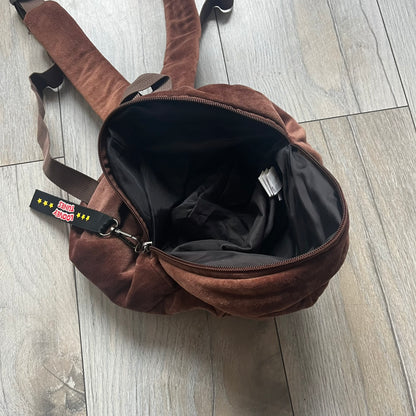 Small plush backpack made from soft brown material, featuring a large 3D Tasmanian Devil face on the front panel. Equipped with a zip closure detailed with a black Looney Tunes branded zipper pull, similar to a keychain tag. Finished with original Looney Tunes tags and compact backpack proportions suitable for light carry.