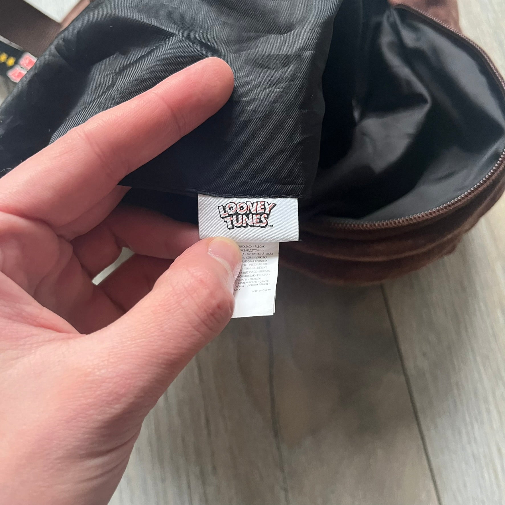 Small plush backpack made from soft brown material, featuring a large 3D Tasmanian Devil face on the front panel. Equipped with a zip closure detailed with a black Looney Tunes branded zipper pull, similar to a keychain tag. Finished with original Looney Tunes tags and compact backpack proportions suitable for light carry.
