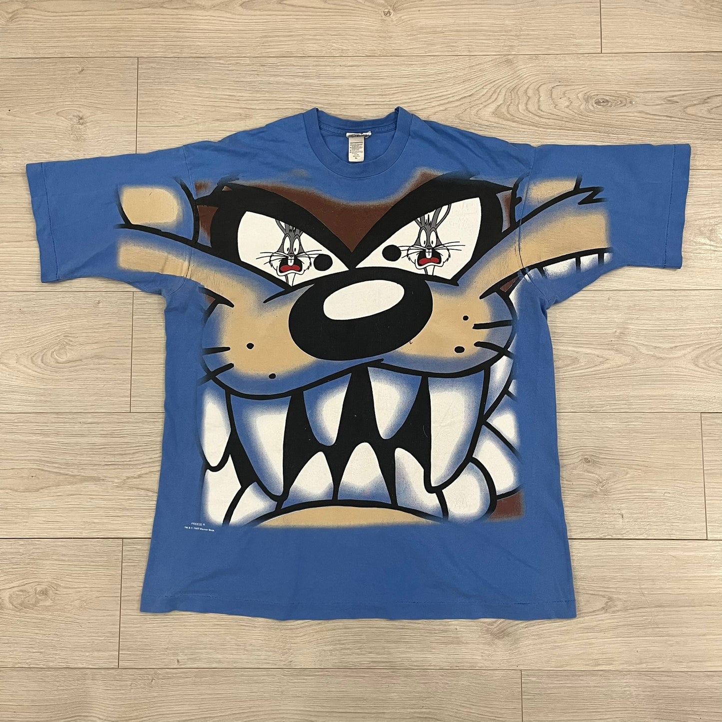 Looney Tunes “Taz & Bugs Bunny” 1997 Freeze Tee featuring a blue base with an all-over front print. The graphic shows Taz with an angry expression and Bugs Bunny reflected in his eyes, with 1997 Warner Bros. Freeze licensing printed below. Made in the USA from 100% cotton.