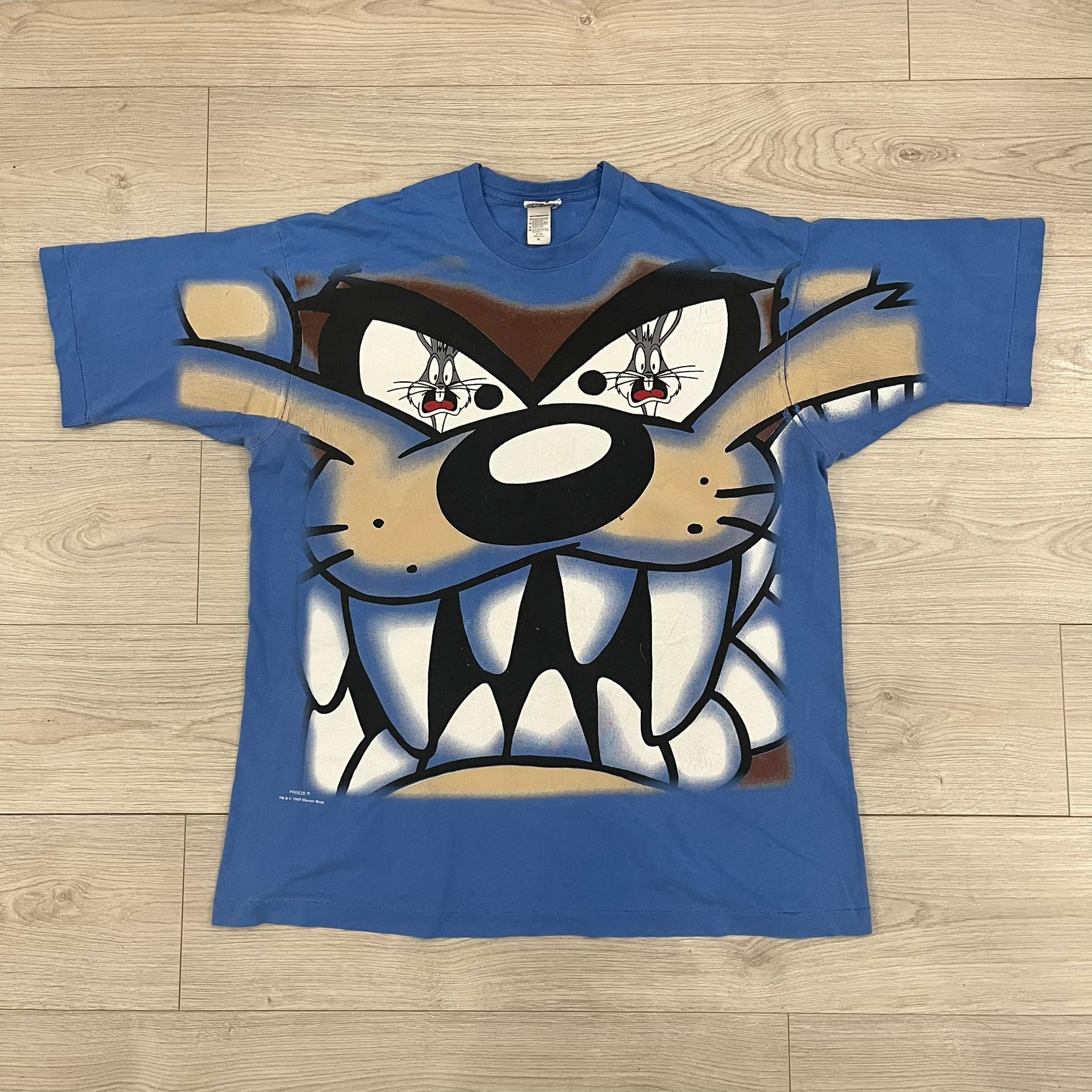 Looney Tunes “Taz & Bugs Bunny” 1997 Freeze Tee featuring a blue base with an all-over front print. The graphic shows Taz with an angry expression and Bugs Bunny reflected in his eyes, with 1997 Warner Bros. Freeze licensing printed below. Made in the USA from 100% cotton.