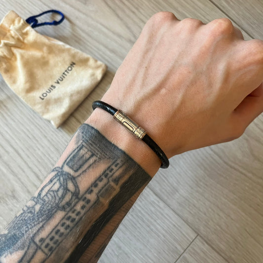 Hand wearing a black and gold bracelet with a Louis Vuitton pouch in the background.