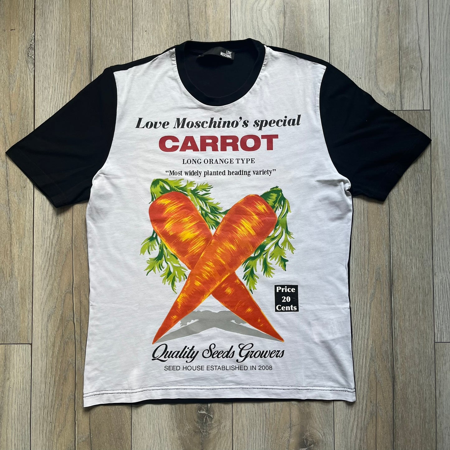Love Moschino 2010s Carrot Tee is a black and white T-shirt with black sleeves and back panel. The front features Love Moschino branding above two illustrated carrots, with large red “CARROT” text underneath and “Price 20 Cents” text below. The front graphic is designed in the style of a vintage carrot advertisement.
