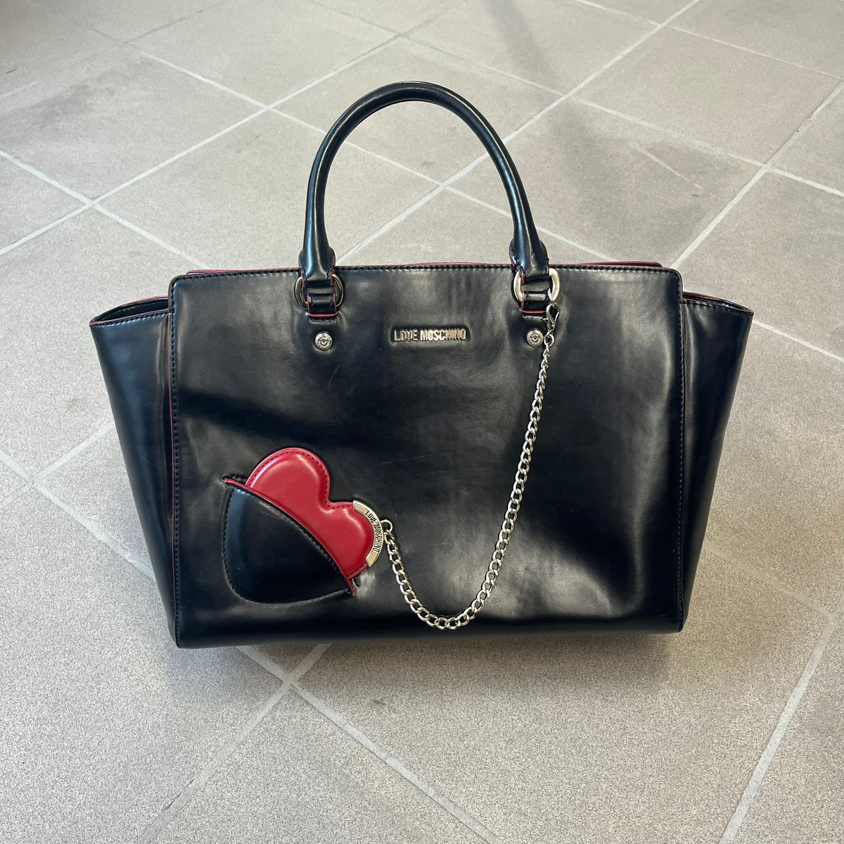Love Moschino FW16 Heart Mirror Black Handbag is a sturdy tote-style handbag in black leather-look material with a red interior lining, two handles, a metal Love Moschino logo on the front, and branded buttons. It features a front pocket holding a heart-shaped mirror with a red back, attached to one handle by a chain.