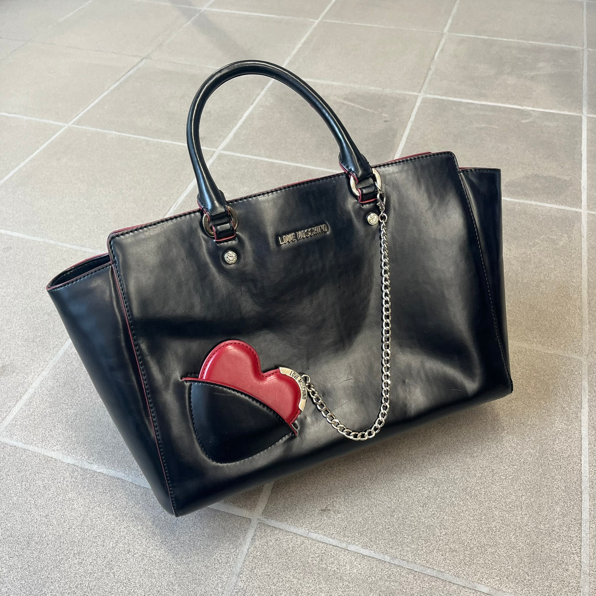 Love Moschino FW16 Heart Mirror Black Handbag is a sturdy tote-style handbag in black leather-look material with a red interior lining, two handles, a metal Love Moschino logo on the front, and branded buttons. It features a front pocket holding a heart-shaped mirror with a red back, attached to one handle by a chain.