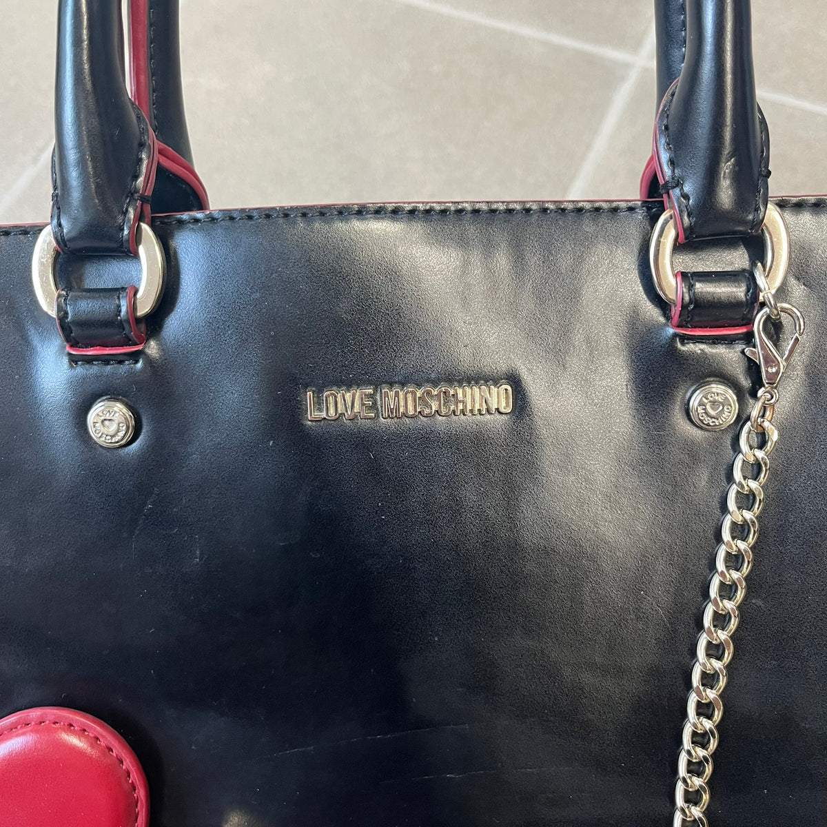Love Moschino FW16 Heart Mirror Black Handbag is a sturdy tote-style handbag in black leather-look material with a red interior lining, two handles, a metal Love Moschino logo on the front, and branded buttons. It features a front pocket holding a heart-shaped mirror with a red back, attached to one handle by a chain.