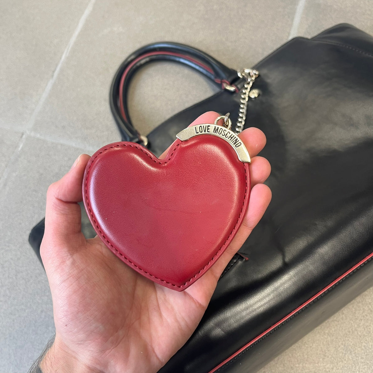 Love Moschino FW16 Heart Mirror Black Handbag is a sturdy tote-style handbag in black leather-look material with a red interior lining, two handles, a metal Love Moschino logo on the front, and branded buttons. It features a front pocket holding a heart-shaped mirror with a red back, attached to one handle by a chain.