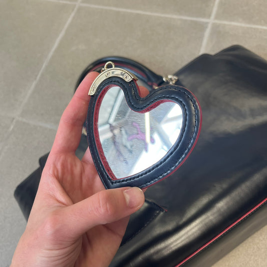 Love Moschino FW16 Heart Mirror Black Handbag is a sturdy tote-style handbag in black leather-look material with a red interior lining, two handles, a metal Love Moschino logo on the front, and branded buttons. It features a front pocket holding a heart-shaped mirror with a red back, attached to one handle by a chain.